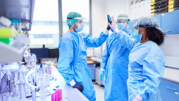 Tres científicos con equipo de protección personal completo, que incluye batas, cubrebocas y protectores faciales, se dan un choque de manos en un laboratorio.